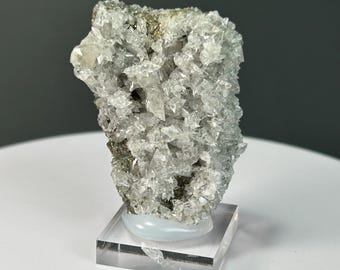 May include: A cluster of clear, crystalline mineral formations with a rough, textured surface. The mineral sits atop a clear, rectangular acrylic base with a white, curved support. The mineral has areas of gold and dark grey.