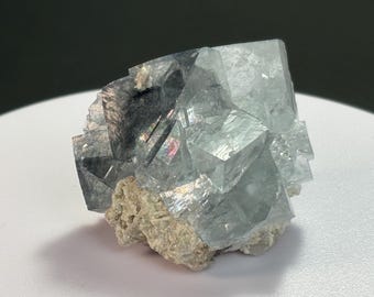 May include: A close-up of a fluorite crystal cluster. The crystal is a pale blue-green color with a cubic structure. The base is a rough, beige-colored matrix. The crystal has a reflective, glassy appearance.