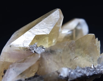 May include: Close-up of a cluster of translucent, pale yellow calcite crystals. The crystals have sharp, pointed terminations and a slightly rough texture. The background is a dark, blurred gradient, highlighting the crystals' details.