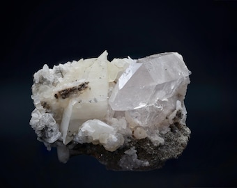 May include: A cluster of clear quartz crystals with a large, faceted central point. The quartz is set against a dark background, with smaller, opaque white crystals and dark gray matrix. The overall effect is a display of natural mineral beauty.