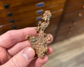May include: A raw copper specimen with a rough, textured surface and a mix of brown and green hues. The natural mineral formation is held in a hand, showcasing its irregular shape and metallic luster. The background features a wooden cabinet.