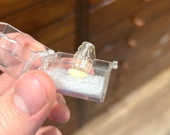 May include: A clear, rectangular plastic case holding a rough, translucent crystal and a small, yellow, oval-shaped stone. The crystal has a jagged, irregular shape. The case rests on a bed of white material.