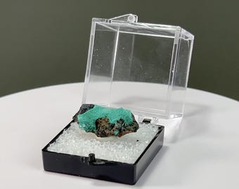 May include: A vibrant, teal-colored mineral specimen displayed in a clear, hinged plastic case. The mineral rests on a bed of small, white, crystalline material within a black base. The case is open, revealing the specimen.