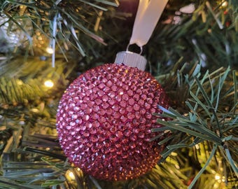 May include: A pink sparkly Christmas ornament hanging on a green artificial Christmas tree with white lights.