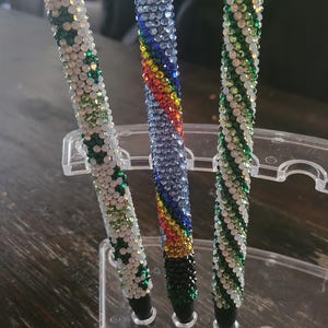 May include: Three pens with a sparkly, crystal-like finish. The pens are displayed in a clear plastic holder. One pen is blue and rainbow, one is green and white, and one is green and white with a hint of gold.