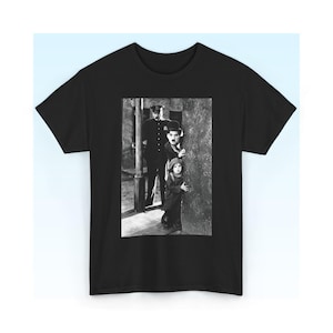 May include: A black short-sleeved t-shirt featuring a rectangular black and white photographic print on the chest. The image depicts a scene with a police officer standing behind Charlie Chaplin and a child who are peeking around a corner.