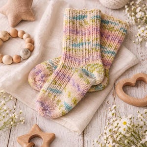 May include: A pair of hand-knitted baby socks in pastel colors of pink, blue, and green, resting on a cream-colored cloth. The image also includes a wooden star, a wooden heart, a wooden bead bracelet, and baby booties.