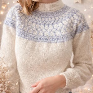 Könnte beinhalten: Cremefarbener Strickpullover mit hellblauem geometrischen Muster auf Brust und Schultern. Der Pullover hat einen gerippten Ausschnitt und Bündchen. Das Model trägt eine blaue Jeans. Der Hintergrund ist weich verschwommen mit warmen, neutralen Tönen.