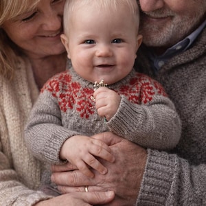 Peut inclure: Un bébé souriant portant un pull gris avec des motifs floraux rouges, tenant un petit bouquet. Le bébé est tenu par deux personnes, probablement des grands-parents, dans un cadre chaleureux et doucement éclairé. Le pull est en tricot douillet.