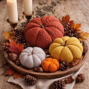 Peut inclure: Une composition automnale sur un plateau en bois. Elle comprend quatre citrouilles tricotées orange, jaune et grise, entourées de pommes de pin, de glands et de feuilles d'automne. Deux bougies allumées dans des bougeoirs en laiton sont en arrière-plan.