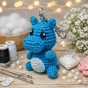 Peut inclure: Un porte-clés hippopotame bleu crocheté à la main avec un ventre blanc. L'hippopotame a des yeux brodés noirs et une chaîne argentée. Le fond comprend des fournitures de couture, des perles et un coussin à épingles.