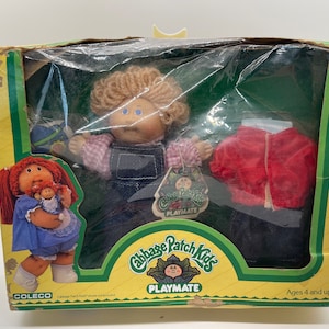 May include: A Cabbage Patch Kids Playmate doll with blonde hair and blue eyes wearing overalls and a plaid shirt. The doll is holding a ball. The doll is packaged in a box with a red and blue design. The box also includes a red jacket and a pair of blue jeans.
