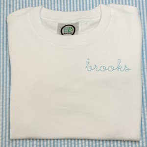 May include: White long-sleeve shirt with a blue embroidered name "brooks" on the front. The shirt has a small green label with the letters "BB" and the text "Bende Boutique" on the front.