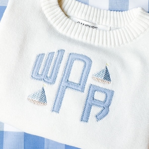 May include: White knitted sweater with a blue embroidered monogram "WPR" and two small blue sailboats. The sweater has a ribbed neckline and is made by the brand "Campure".