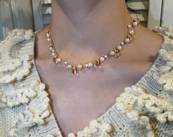 May include: A delicate gold necklace featuring small white pearls and pink teardrop-shaped beads. The necklace is worn against a cream-colored sweater.