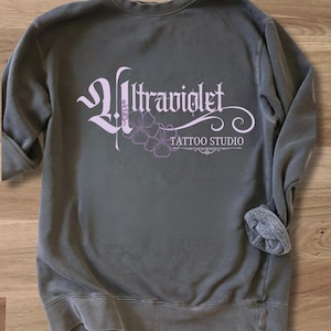 May include: A gray sweatshirt with the text "Ultraviolet Tattoo Studio" in a light purple script and floral design. The sweatshirt has rolled-up sleeves and a relaxed fit, perfect for casual wear.