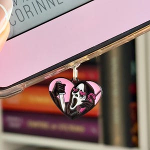 May include: A pink heart-shaped phone charm featuring a black and white illustration of Ghostface from the Scream movie franchise. Ghostface is holding a knife in one hand and a phone in the other. He has a white mask with a scream expression and pink hearts for eyes.