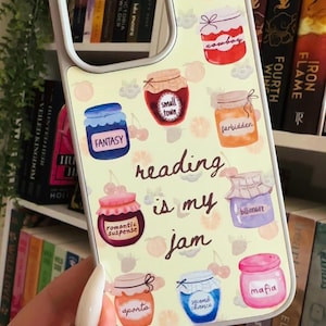 May include: A phone case with a light beige background featuring watercolor illustrations of jam jars with book genres like "Fantasy" and "Mafia." The phrase "reading is my jam" is written in a script font.