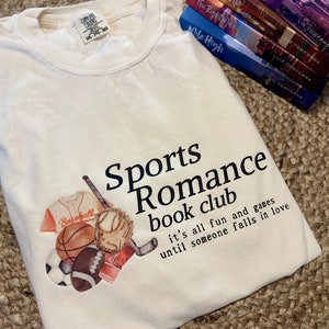 May include: A white t-shirt with a graphic of sports equipment and the text "Sports Romance book club it's all fun and games until someone falls in love".