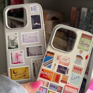 May include: Two white iPhone cases with different designs. The left case has a collage of colorful stamps with text like "Terrasen", "Yvelia", "Arettia", and "Basgiath". The right case has a collage of colorful matchbox labels with text like "Cowboy Romance", "Slow Burn", "Forced Romance", "Small Town", and "Single".