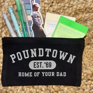 May include: Black canvas pouch with white text that reads "POUNDTOWN EST. '69 HOME OF YOUR DAD". The pouch is filled with various items, including a sticker of a man in a suit, a pack of transparent sticky notes, and several blue and green pens.