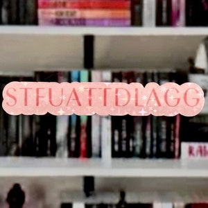 May include: A pink and white sticker with the text "STFUATTIDLAGG" on a bookshelf with books in the background.