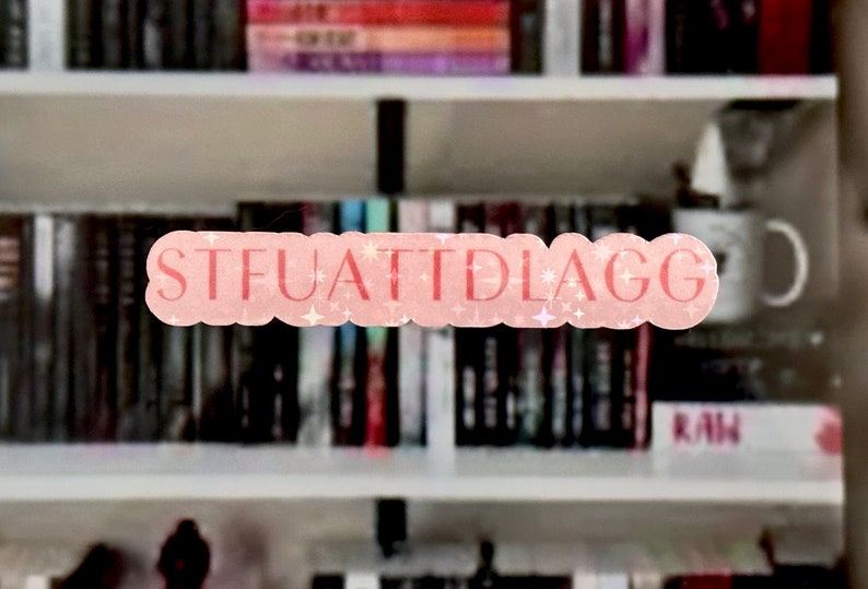 May include: A pink and white sticker with the text "STFUATTIDLAGG" on a bookshelf with books in the background.