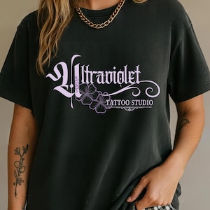 May include: Dark gray t-shirt with the text "Ultraviolet Tattoo Studio" in a stylized, light purple font. The design includes floral elements. The shirt is worn by a person.