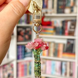 May include: A gold-toned keychain with a heart-shaped charm and a small bouquet of pink and red flowers. The bouquet is made of beads and wire, with green beads forming the stems. The keychain is held up by a gold clasp.