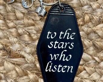 May include: Black motel key chain with the text "to the stars who listen" in white lettering. The keychain has a silver ring with a silver dragon charm and a silver book charm.