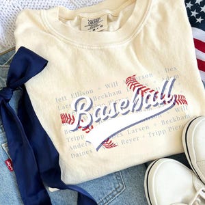 May include: Cream-colored baseball-themed sweatshirt with the word "Baseball" in a script font, accented with red stitching details. The shirt is paired with blue denim shorts, a navy blue ribbon, and white sneakers.