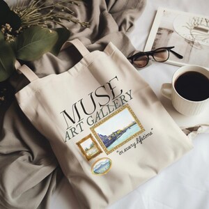 May include: Cream-colored tote bag with the text "MUSE ART GALLERY" and three framed watercolor paintings. The bag has two handles and the phrase "in every lifetime" is printed below the paintings. A cup of coffee and glasses are in the background.