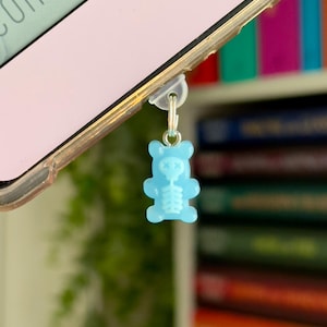 May include: A light blue gummy bear phone charm with a skeleton design. The charm is attached to a phone with a clear case. The background includes a bookshelf with colorful books and a pink phone.