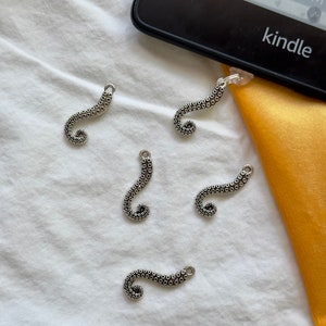 May include: Five silver octopus-shaped bookmark charms on a white fabric background. The charms are all in the shape of an octopus tentacle, with a curled end. The charms are all the same size and shape.