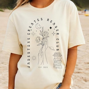May include: Cream-colored t-shirt with a black graphic design. The design features the text "Jadestone Creates Reader Club" in an arch, with illustrations of a hand holding flowers, a stack of books, and celestial elements. The text "EST. 2023" is also included.