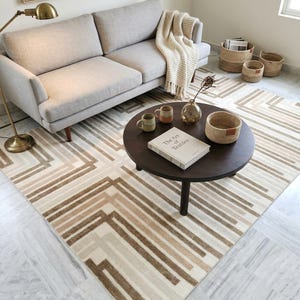May include: A living room scene featuring a light gray sofa, a round dark brown coffee table, and a geometric patterned rug in shades of beige and brown. Woven baskets and a throw blanket add texture. A floor lamp and artwork complete the cozy space.