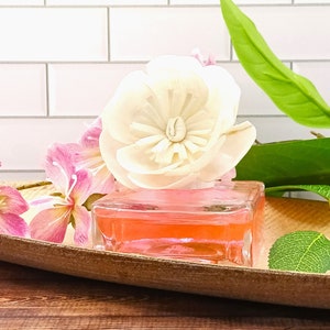 May include: A clear glass diffuser bottle filled with pink liquid and topped with a white flower. The diffuser is placed on a wooden tray with a pink flower and a green leaf.