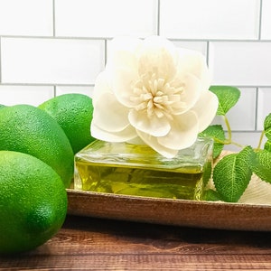 May include: A glass diffuser bottle with a white wooden flower on top. The bottle is filled with a yellow liquid and is sitting on a wooden tray with green limes.