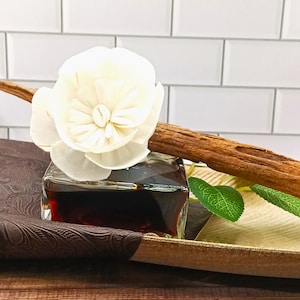 May include: A glass diffuser bottle containing a dark brown liquid, topped with a white wooden flower. The diffuser is placed on a brown tray with a decorative pattern.