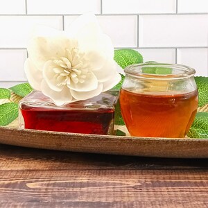 May include: A white wooden flower sits on top of a clear glass bottle filled with a red liquid. Next to it is a clear glass jar filled with a golden liquid. Both bottles are on a wooden tray with green leaves.