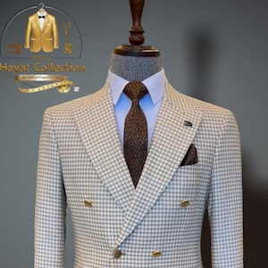 May include: A double-breasted suit in a houndstooth pattern, displayed on a mannequin. The suit features a light gray and white checkered blazer with gold buttons, paired with brown trousers. A blue dress shirt and patterned tie complete the ensemble.