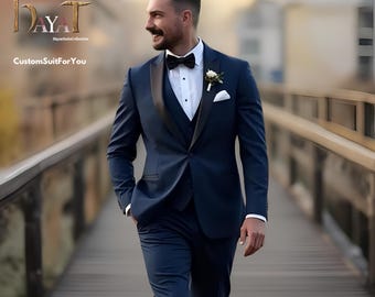 May include: A man wearing a dark blue suit with a black lapel, white shirt, and black bow tie. He is standing on a wooden bridge with a blurred background.