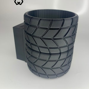 Puede incluir: Portavasos negro impreso en 3D con un diseño de banda de rodadura de neumático. El portavasos tiene una base rectangular y un portavasos cilíndrico.