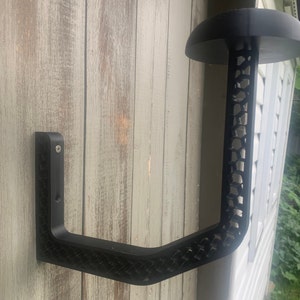 K&ouml;nnte beinhalten: Schwarzer Wandhalter f&uuml;r H&uuml;te aus Metall mit Wabenmuster. Der Halter hat einen gebogenen Arm und eine flache Oberseite zum Aufh&auml;ngen von H&uuml;ten.
