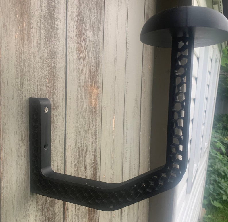 K&ouml;nnte beinhalten: Schwarzer Wandhalter f&uuml;r H&uuml;te aus Metall mit Wabenmuster. Der Halter hat einen gebogenen Arm und eine flache Oberseite zum Aufh&auml;ngen von H&uuml;ten.