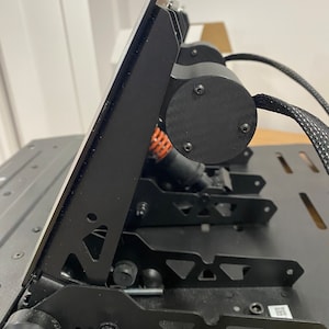 May include: Black metal racing simulator pedals with a silver metal plate on top. The pedals have a triangular design and are connected to a black base.