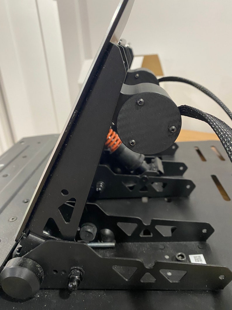 May include: Black metal racing simulator pedals with a silver metal plate on top. The pedals have a triangular design and are connected to a black base.