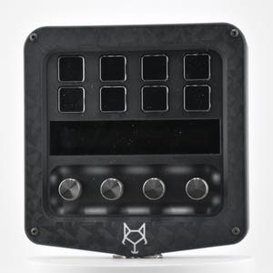 May include: A black, rectangular device with a carbon fiber texture. It has eight square buttons, a rectangular screen, and four round knobs. The device has a white fox logo on the bottom.