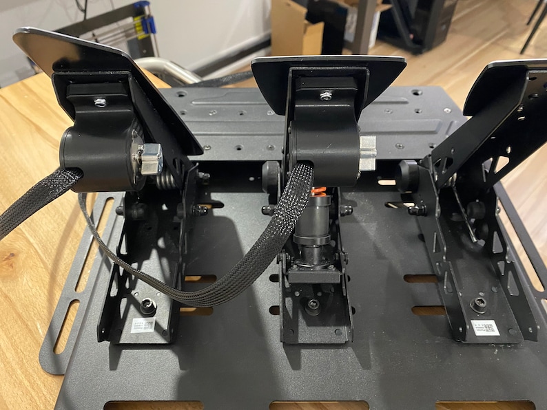 May include: A black metal racing pedal set with three pedals, a brake pedal, a gas pedal, and a clutch pedal. The pedals are connected to a black metal base with a black cable.