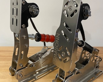 May include: A pair of silver metal racing simulator pedals with a black carbon fiber plate on the left pedal. The pedals are labeled "SIMSOIS".